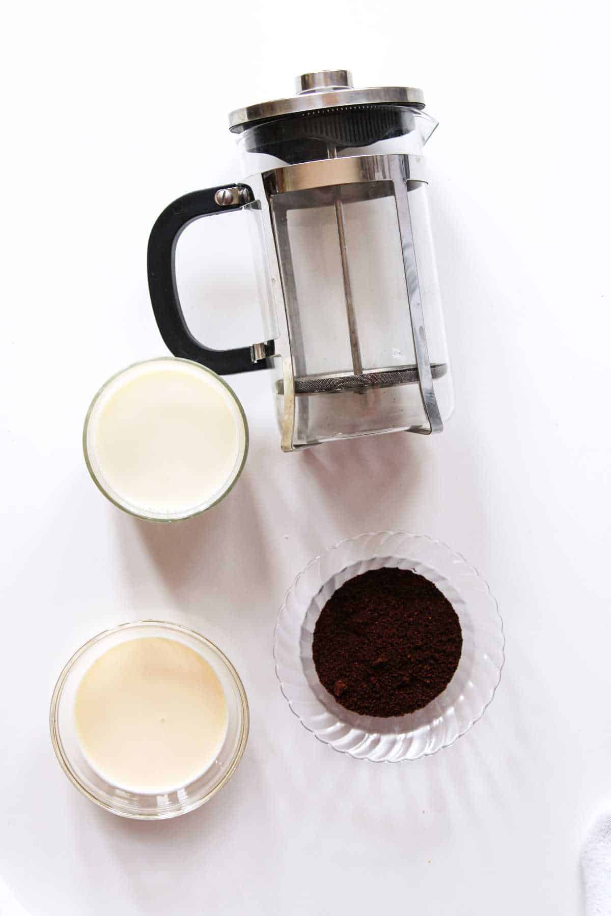 Fremch coffee press with coffee grounds, cream, and milk on a white background.