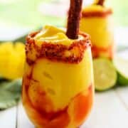 Two Chamoy and Tajin rimmed glasses filled with some mango Mangonada and a tamarind candy stick.