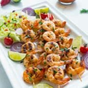 platter of oven shrimp skewers with salad, red onion slices, and lime wedges.
