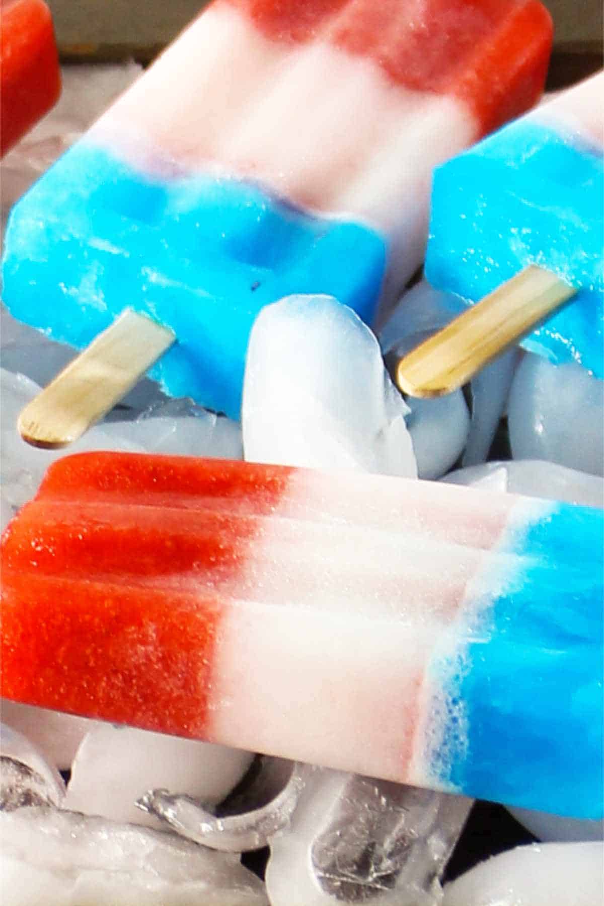 Red, white, and blue popsicles with wooden sticks rest on ice cubes, making these classic red white and blue popsicles the perfect patriotic or summer-themed treat.