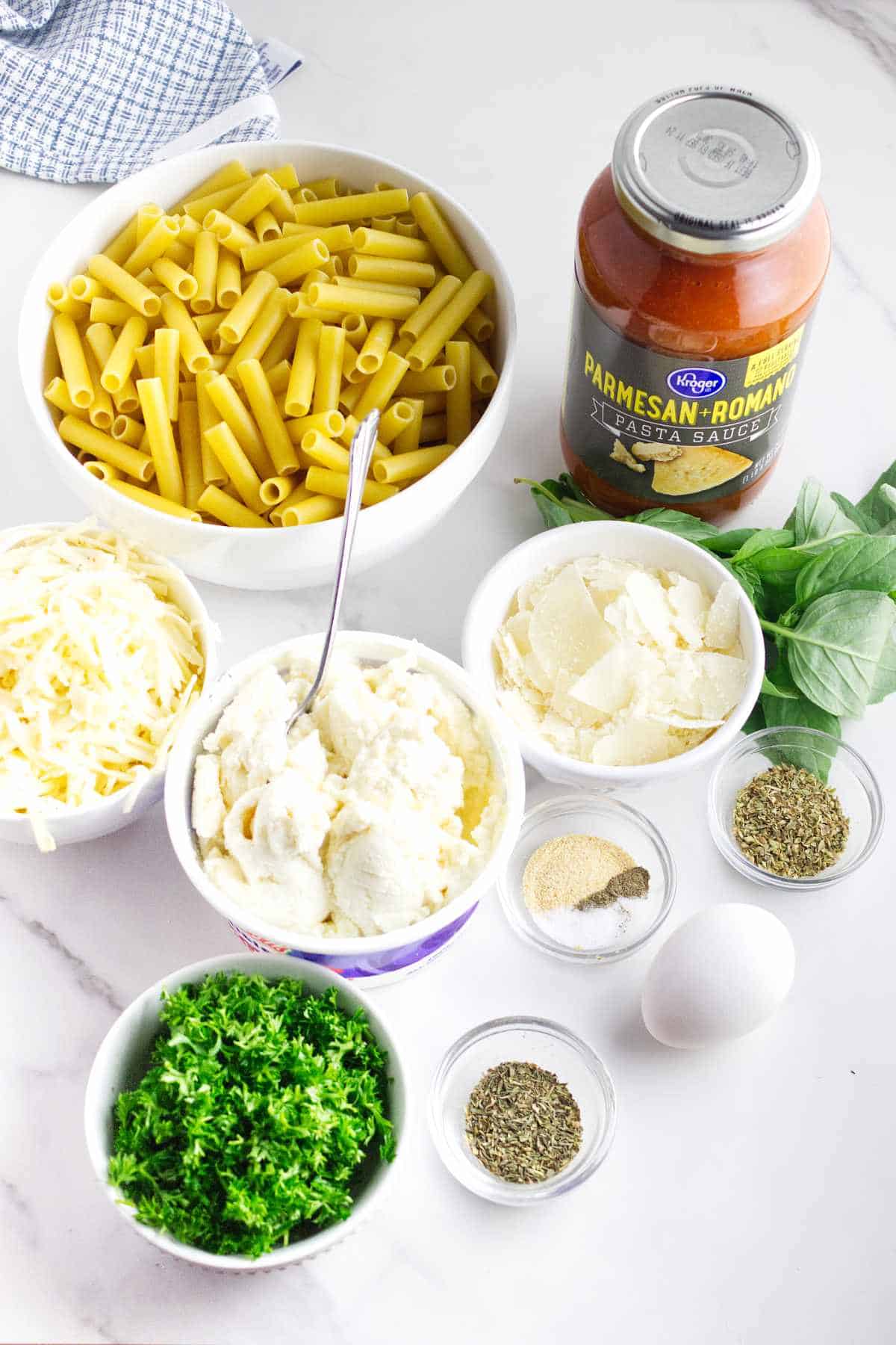 dry pasta, spaghetti sauce, ricotta cheese, shredded mozzarella, grated Parmesan, fresh basil, chopped parsley, an egg, and seasonings on a white background.