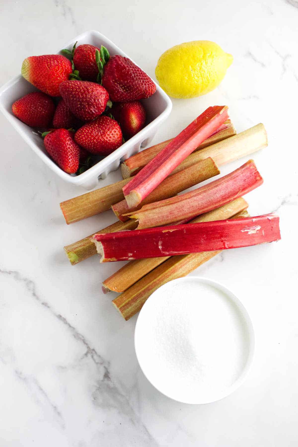 rhubarb stalks, basket of strawberries, sugar, and a lemon on a white background.