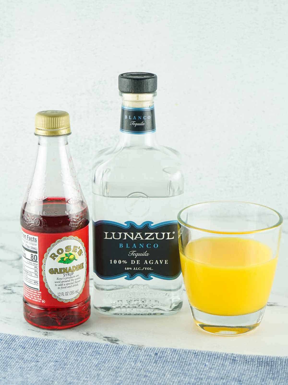 bottle of tequila, grenadine, and a glass or orange juice on a white marble background.