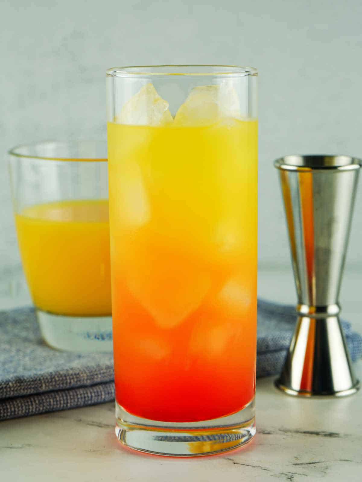 tequila sunrise on a white marble background glass of orange juice and a jigger nearby.