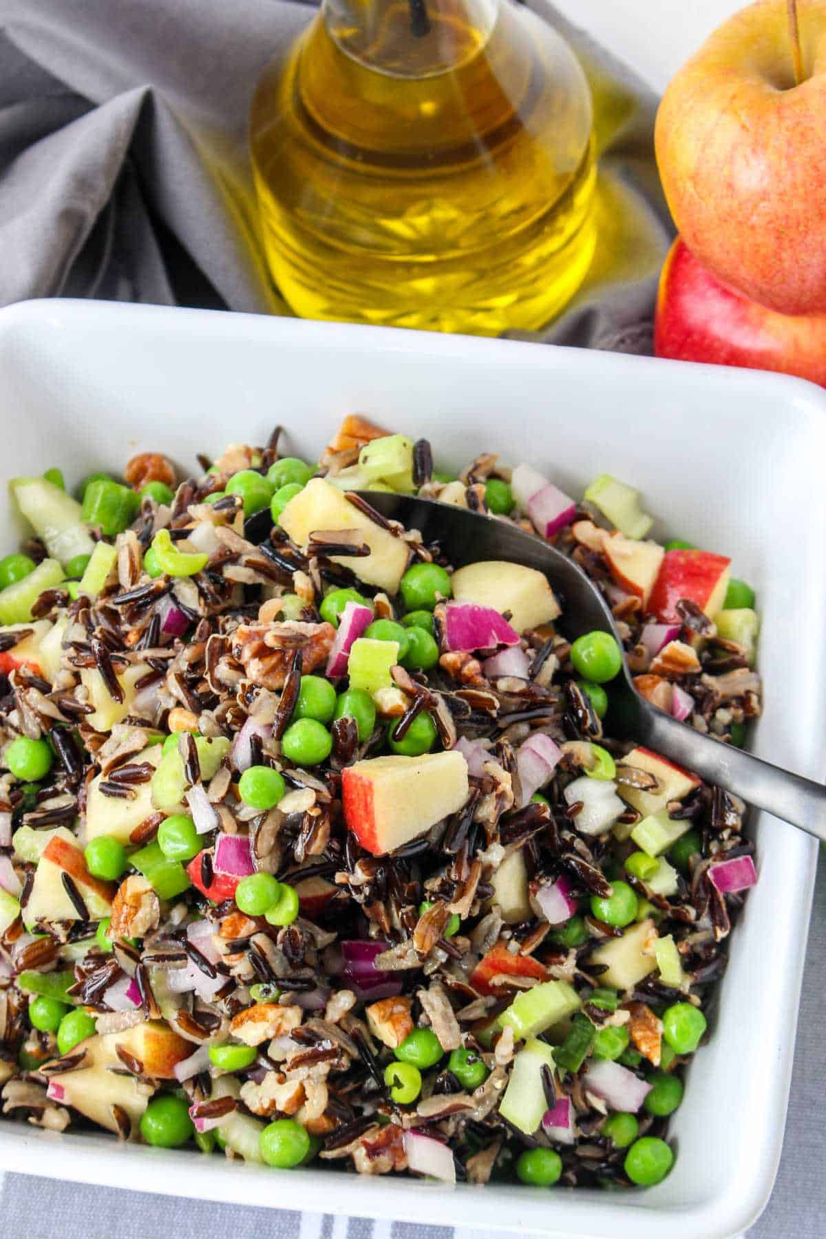 Serving bowl of wild rice salad on a white background with apples and olive oil nearby.