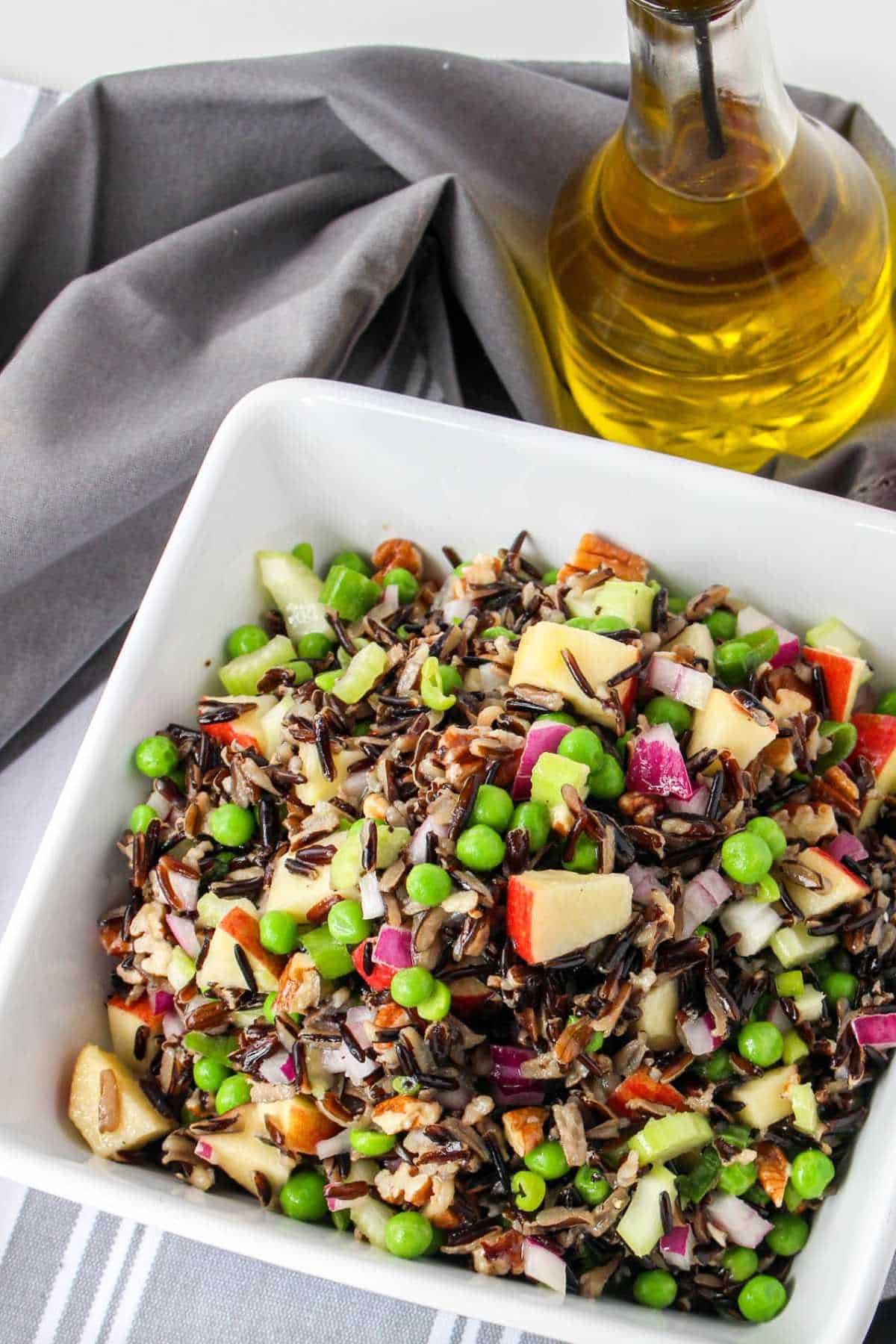 Serving bowl of wild rice salad on a white background with apples and olive oil nearby.