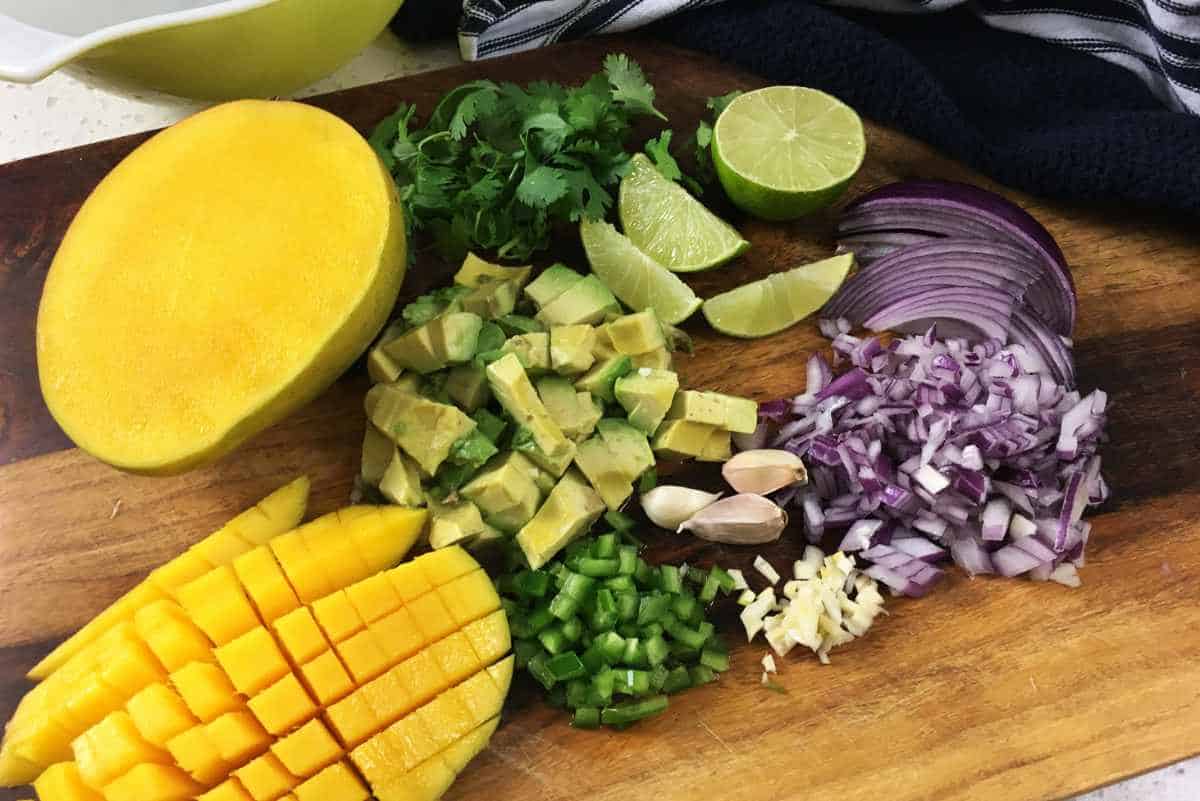 cutting board with chopped red onion, chopped avocado, chopped green bell pepper, and chopped mango, with lime wedges and chopped cilantro.