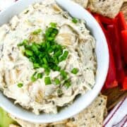 bowl of caramelized onion dip garnished with chives next to radish sliced, crackers, and sliced cucumbers.