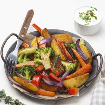 A plate of roasted vegetables including broccoli, carrots, red bell peppers, red onions, and potato wedges, garnished with fresh herbs, with a fork on the side and a small bowl of creamy dipping sauce in the background.
