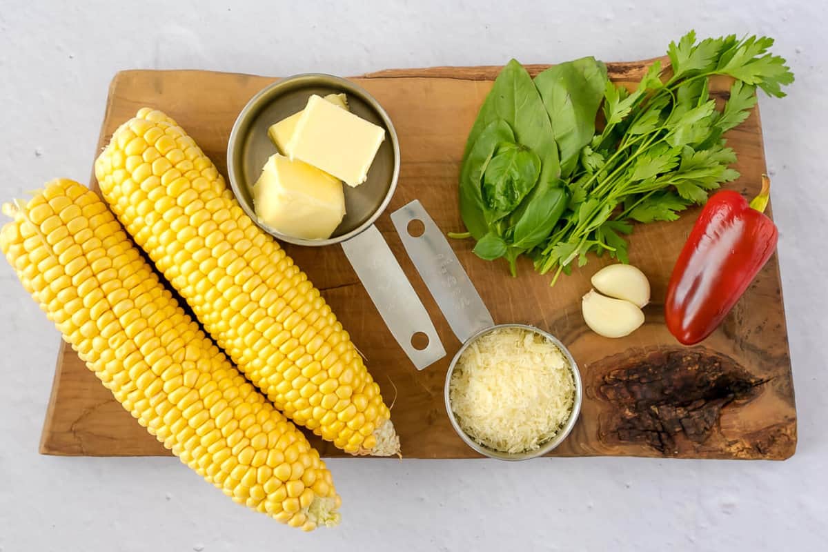 ingredients for making corn on the cob on traeger: ears of corn, grated cheese, herbs, and butter on a wooden cutting board.