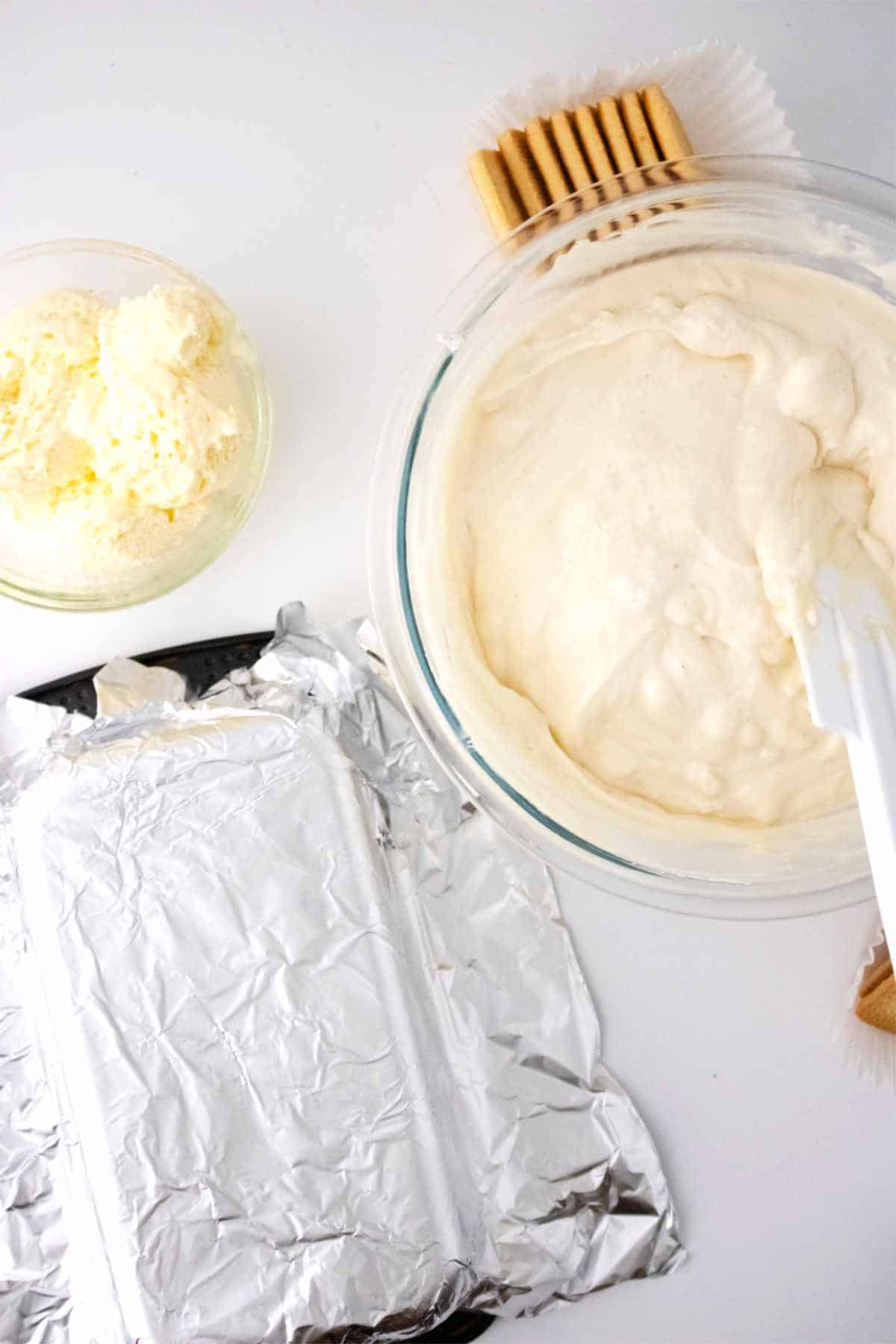cheesecake mixture and whipped ice cram mixture in two bowls with an aluminum foil lined loaf pan nearby.