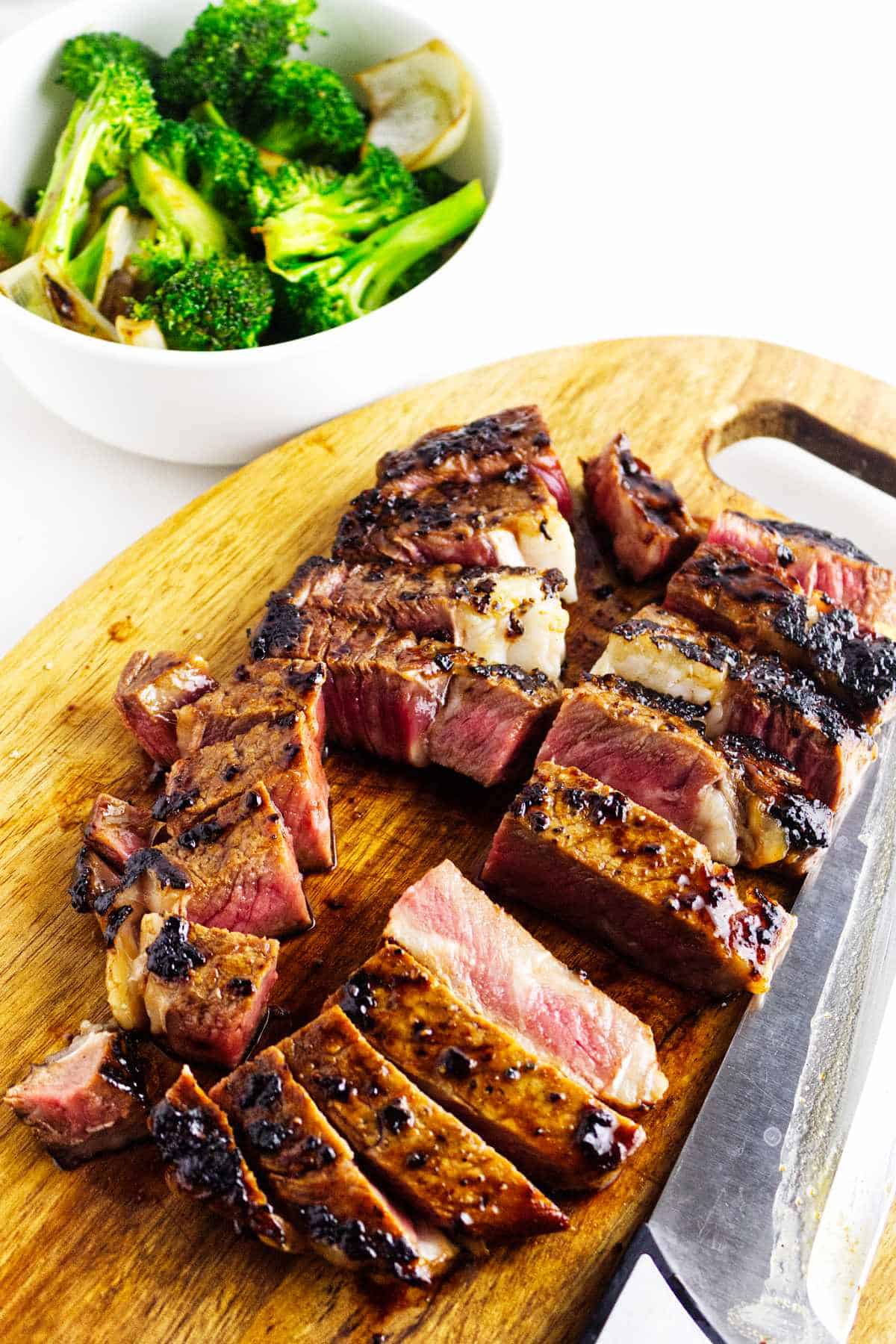 cut grilled beef on a cutting board with a bowl of grilled broccoli in the background.