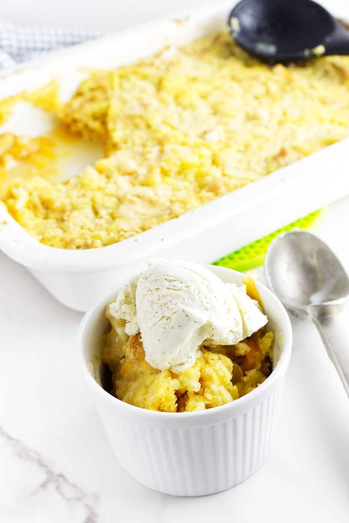 serving of peach dump cake with ice cream on top and baking dish in background.