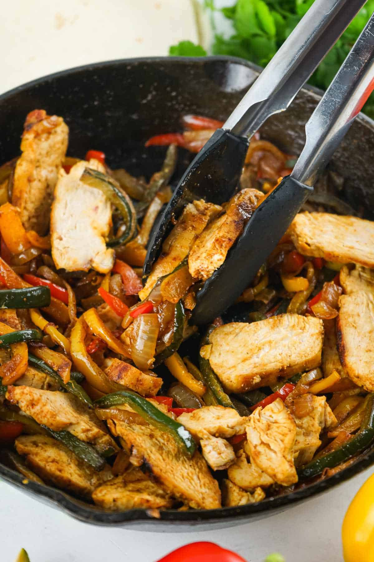 chicken fajitas in cast iron skillet with tongs scooping up portion.
