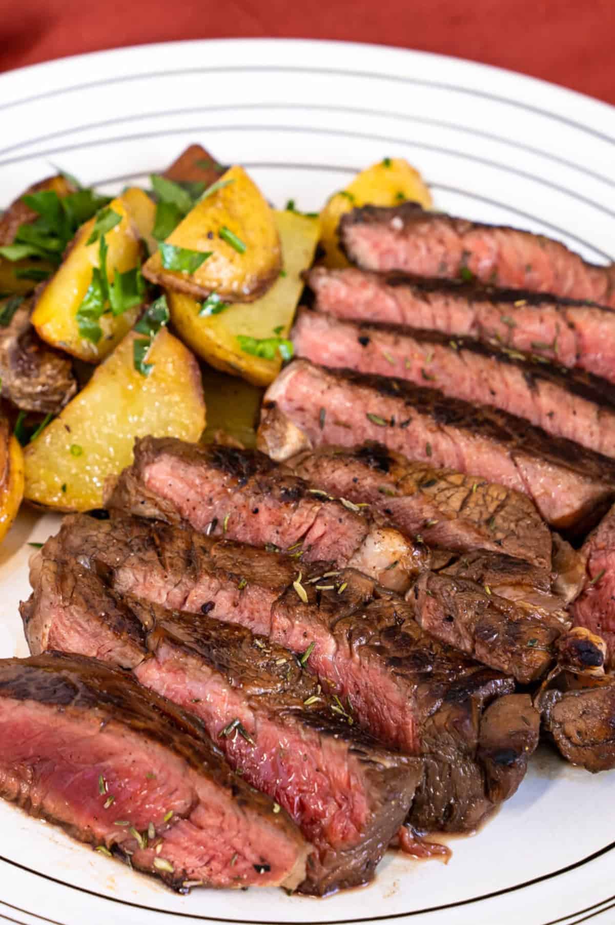 Sliced grilled top sirloin steak, cooked medium-rare with a pink center, is served on a white plate alongside roasted potatoes garnished with fresh herbs.