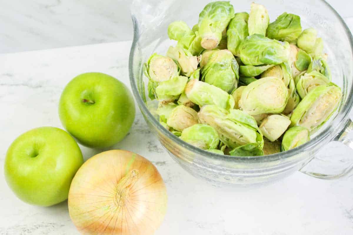 bowl of sliced brussels sprouts and granny smith apples and an onion on a marble background.
