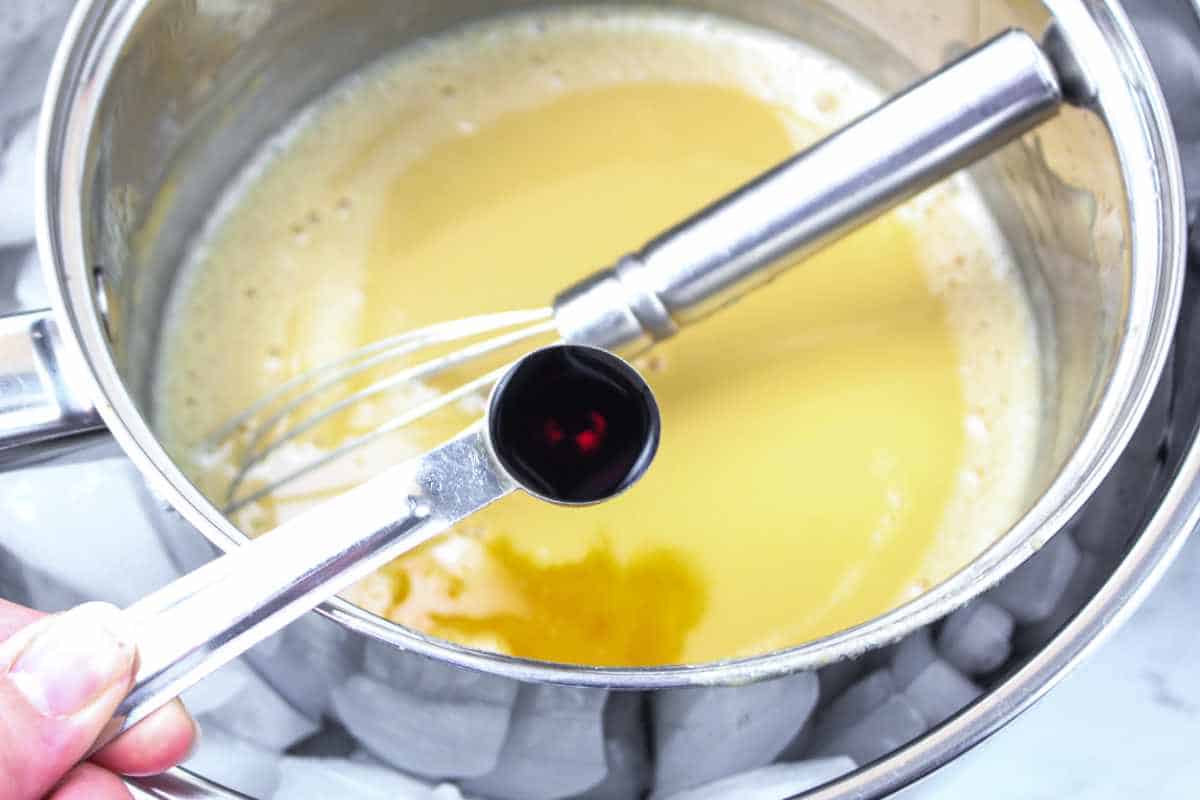 A hand holds a measuring spoon with vanilla extract above a mixing bowl of yellow batter for peach ice cream, with a whisk inside. The bowl sits in a larger bowl filled with ice cubes.