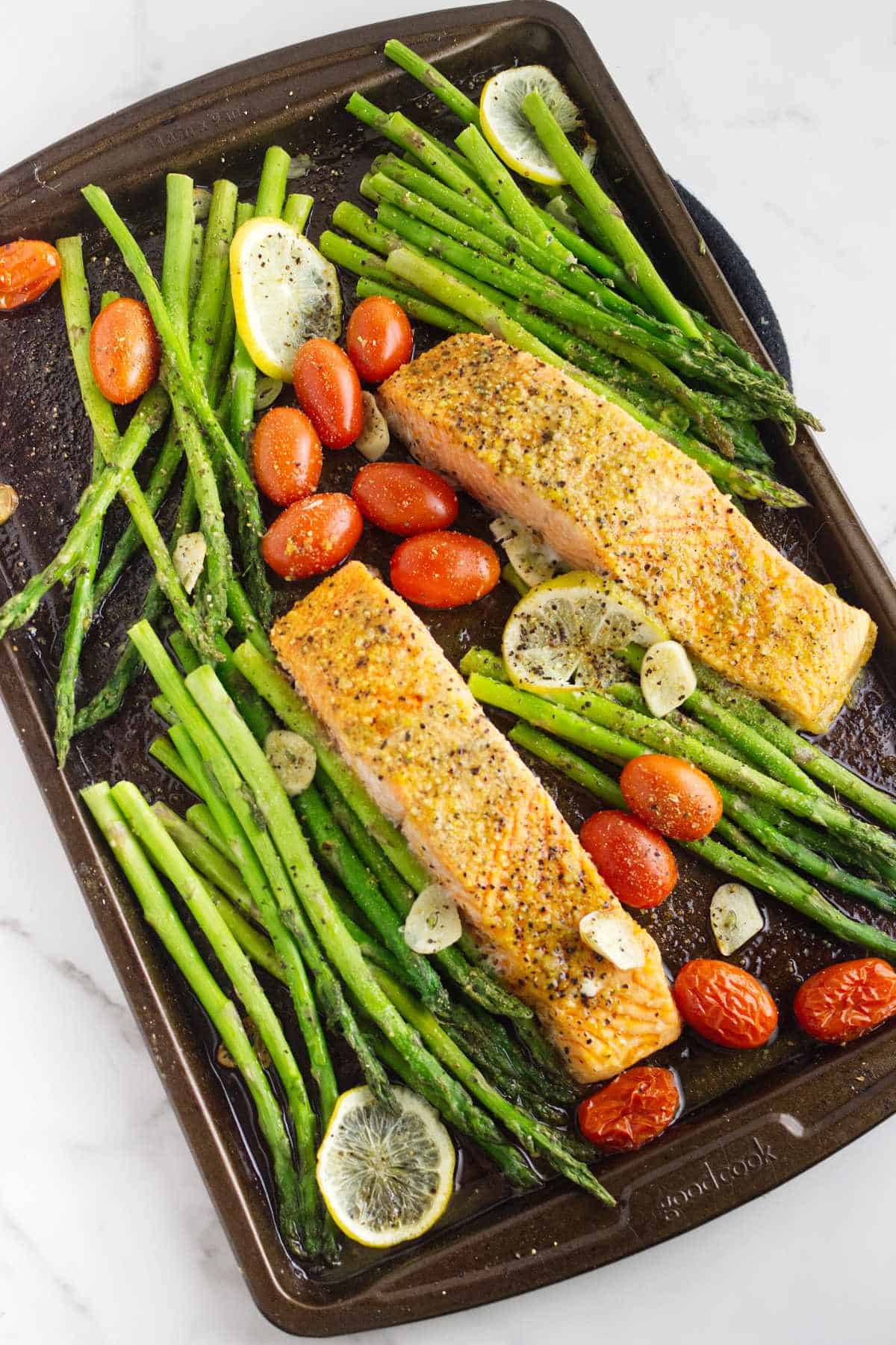 baking tray with roasted seasoned asparagus spears, sliced lemons, blistered tomatoes, and broiled fish filets.