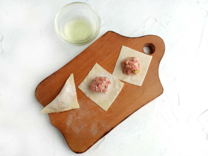 Wooden board with two meat-filled wonton wrappers, one folded wonton, and a small glass bowl of water on a white surface.