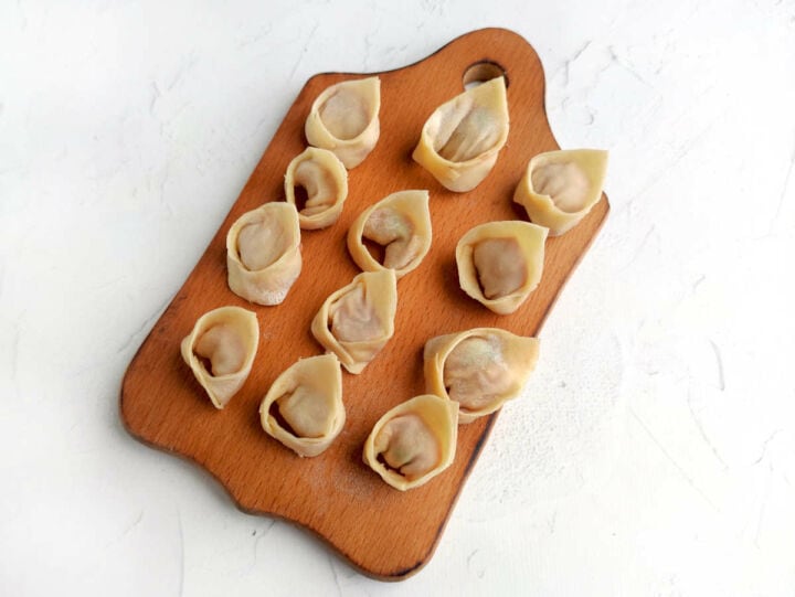 Twelve uncooked dumplings sit neatly on a handled wooden cutting board atop a white textured surface.