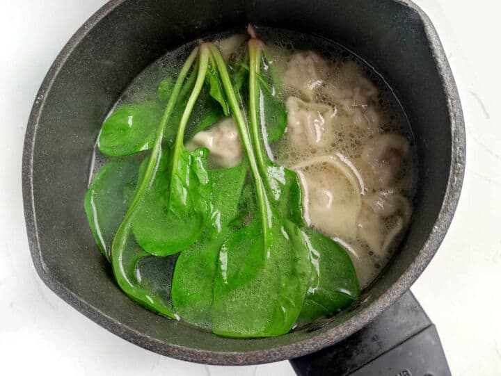Black pot of subgum wonton soup with green spinach and dumplings in clear broth.