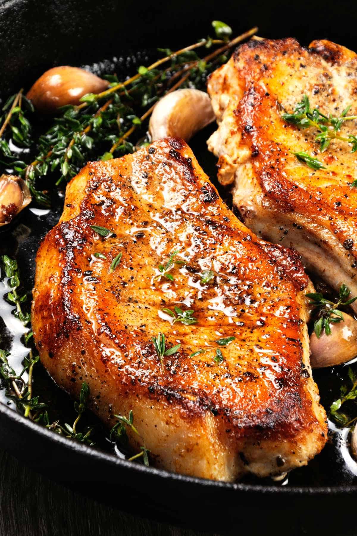Juicy, pan grilled pork chops on a bone in oil with garlic and herbs in a frying pan.