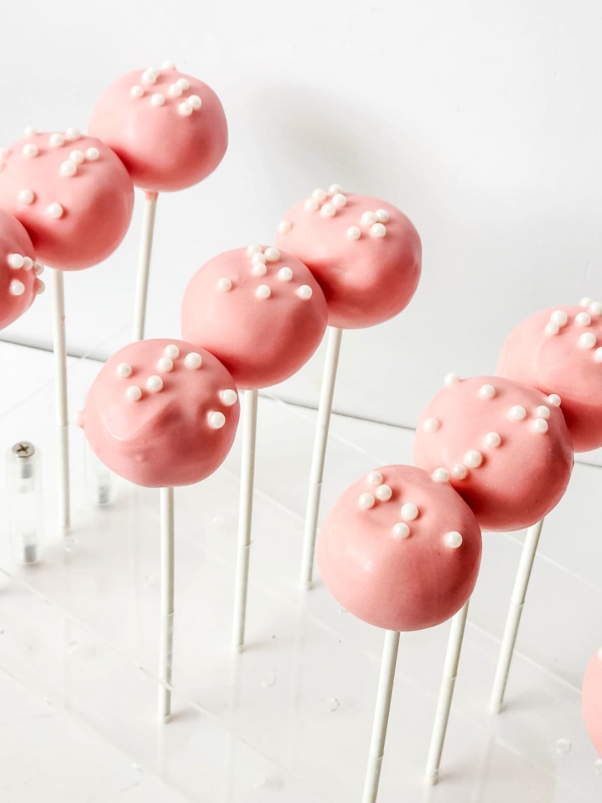 pink cake pops in drying rack with white sprinkles on top.