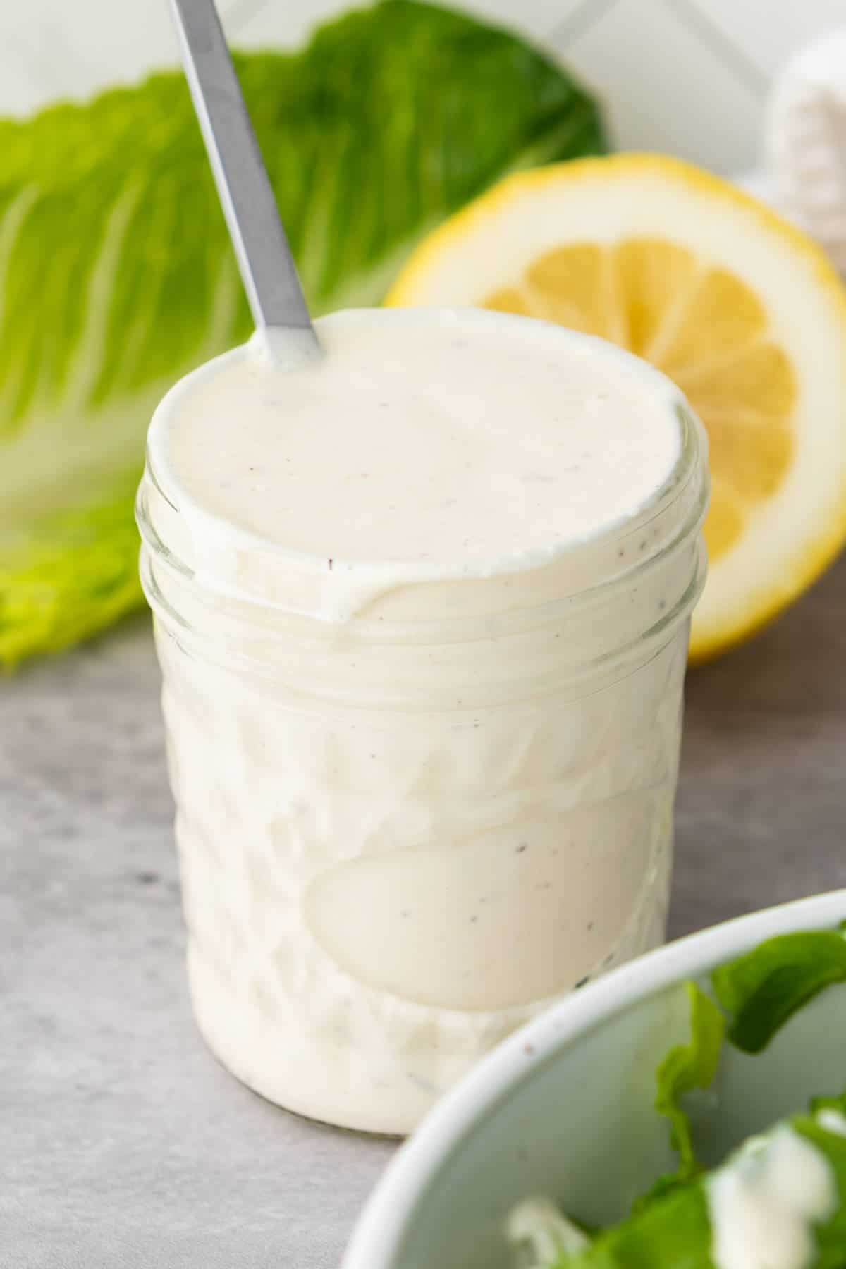 Jar of homemade Caesar salad dressing with a salad in the background.