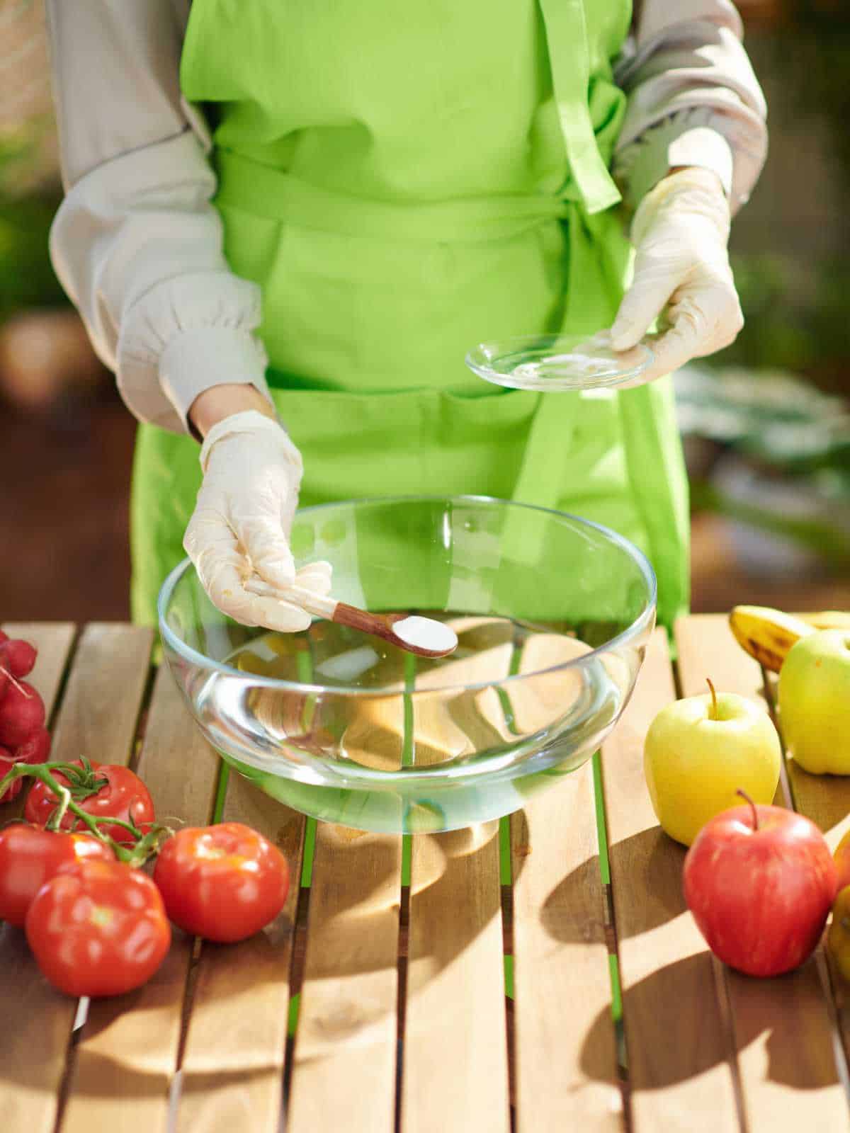 Closeup on housewife in green apron removes pesticides by soaking vegetables and fruits in water with baking soda in modern kitchen.