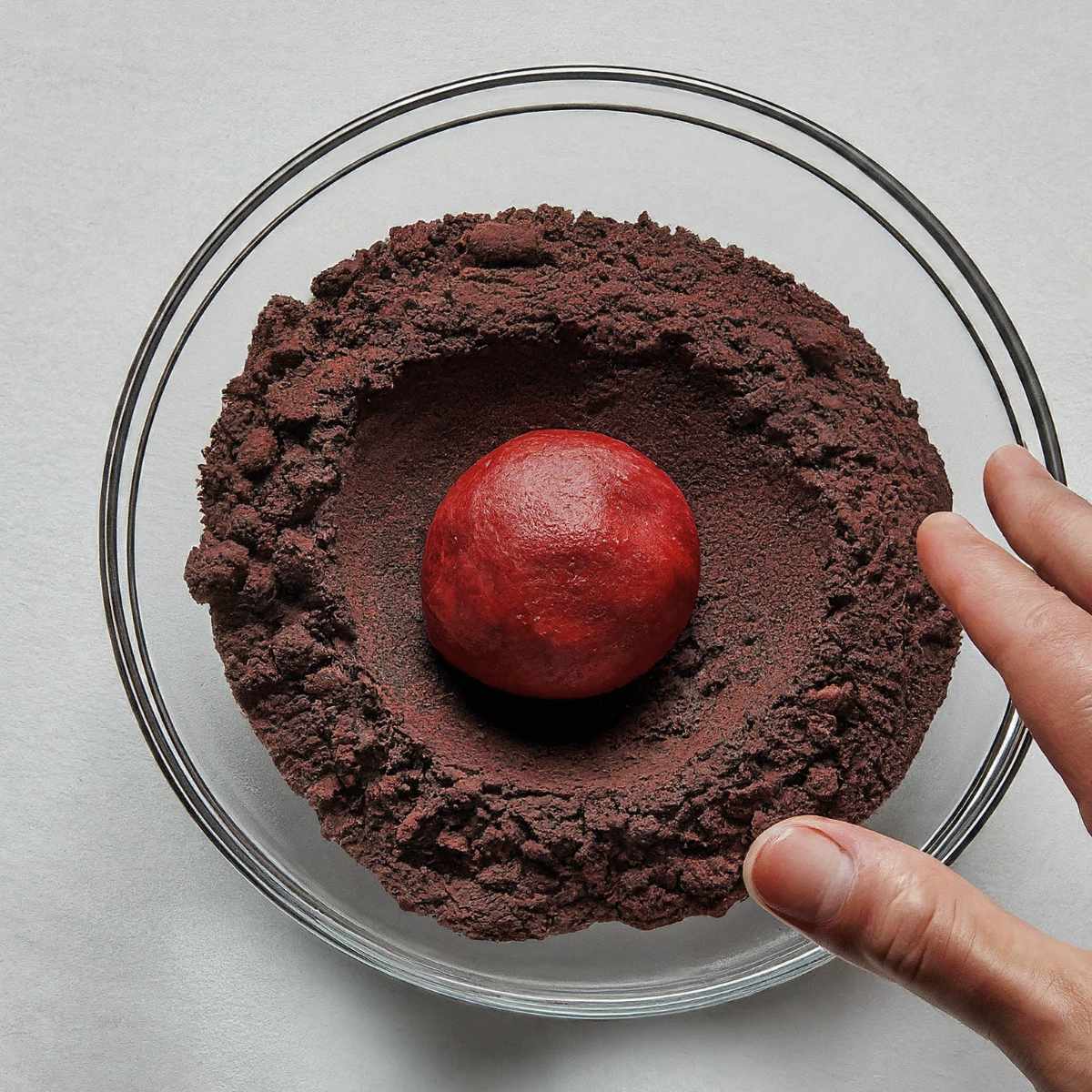 bowl of crushed chocolate Oreo cookies with a ball of red velvet cookie dough being rolled.