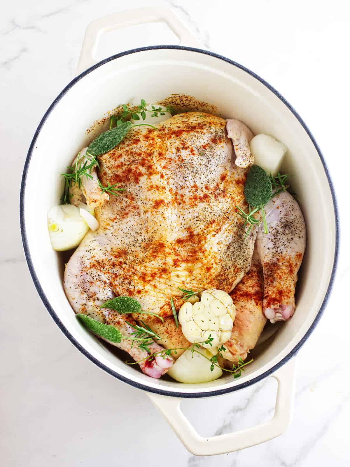 Dutch oven with seasoned raw chicken, garlic bulb, and fresh herbs.