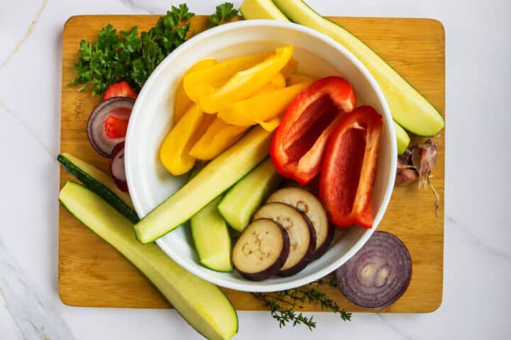 Bowl of sliced zucchini, bell peppers, red onion, and eggplant on a wooden board.