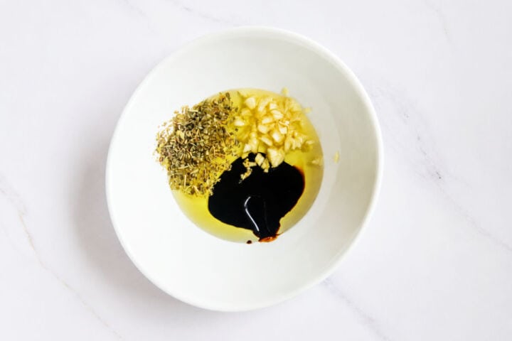 White bowl with olive oil, garlic, dried herbs, and balsamic vinegar on a white marble surface.