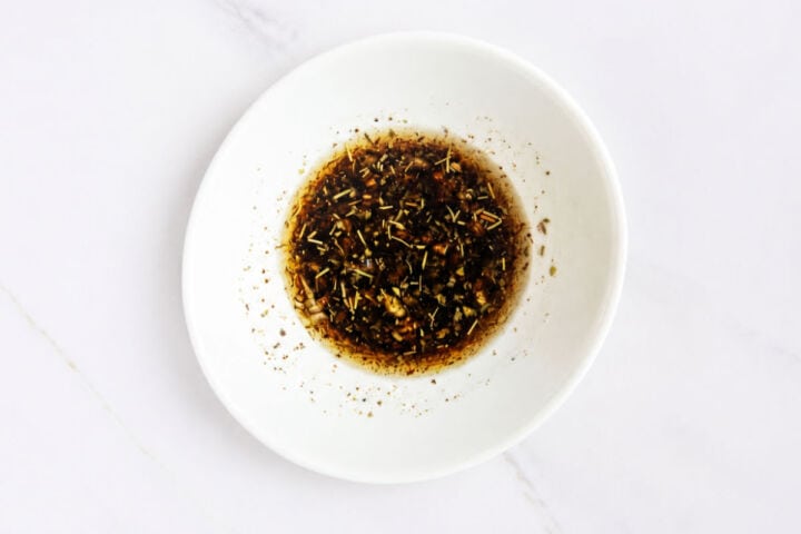 Mixed herb vinegar marinade in a white bowl on a white marble surface.