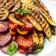Grilled Mediterranean veggies-zucchini, bell peppers, onions-on a white plate with parsley, showing char and grill marks.