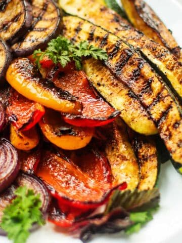 Grilled Mediterranean veggies-zucchini, bell peppers, onions-on a white plate with parsley, showing char and grill marks.