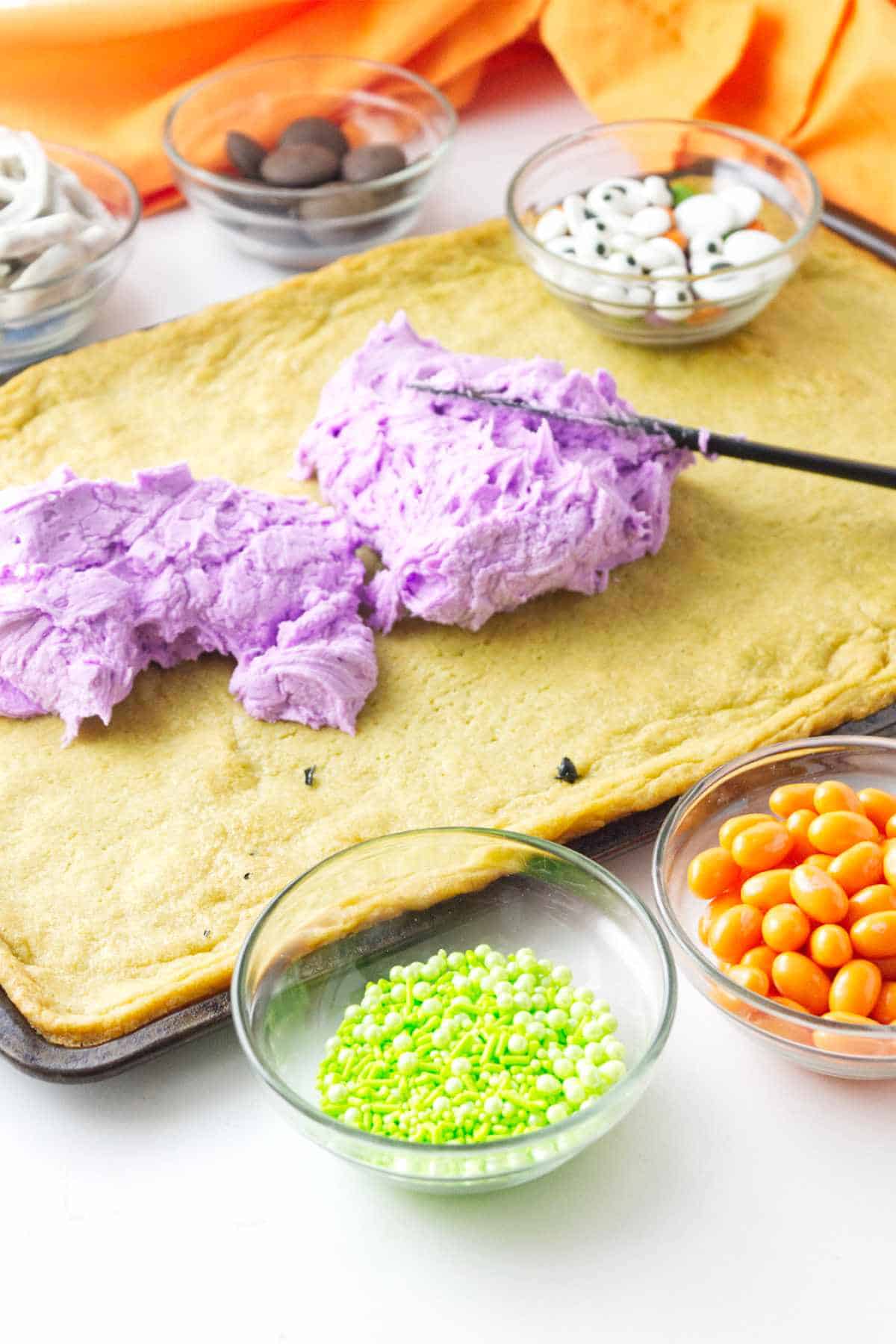 Frosting piles on a baked sheet pan of cookie dough with bowls of decorations nearby.