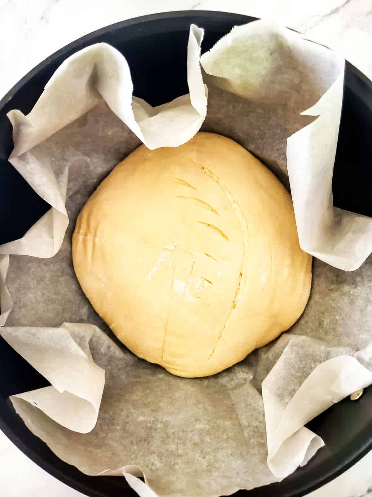 Scored bread dough on parchment paper lined heavy pot..