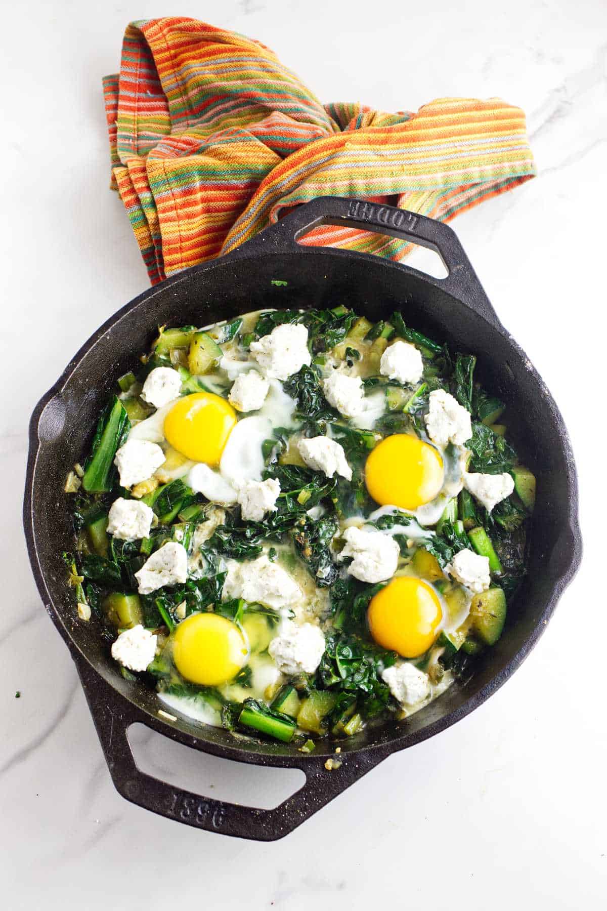 Eggs cracked on top of simmering greens in a skillet. Goat cheese added in between the eggs.