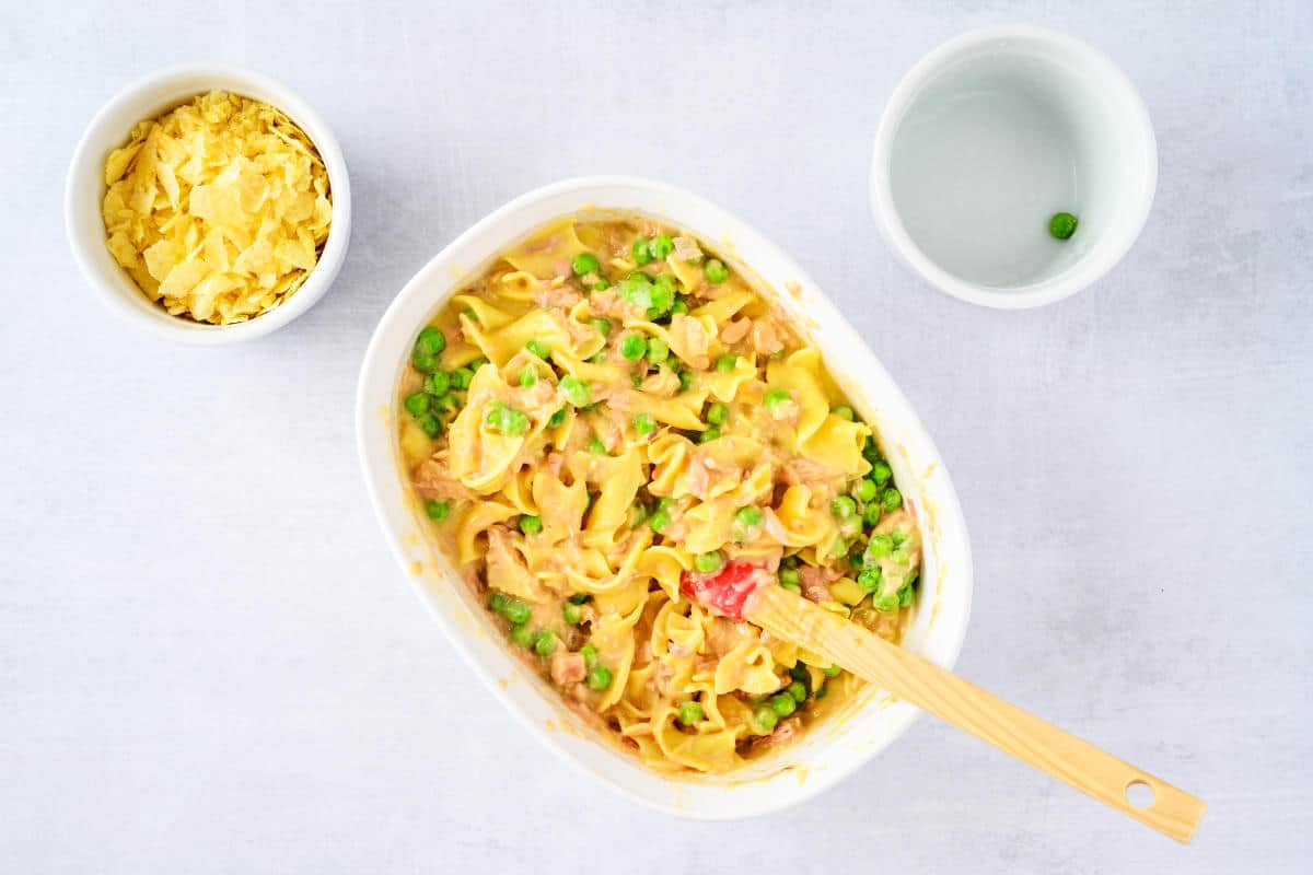 Green peas stirred into tuna casserole, with shredded cheese nearby.