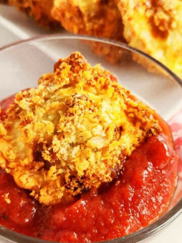 A crispy, golden-brown air fryer toasted ravioli sits in a small glass bowl filled with red marinara sauce.