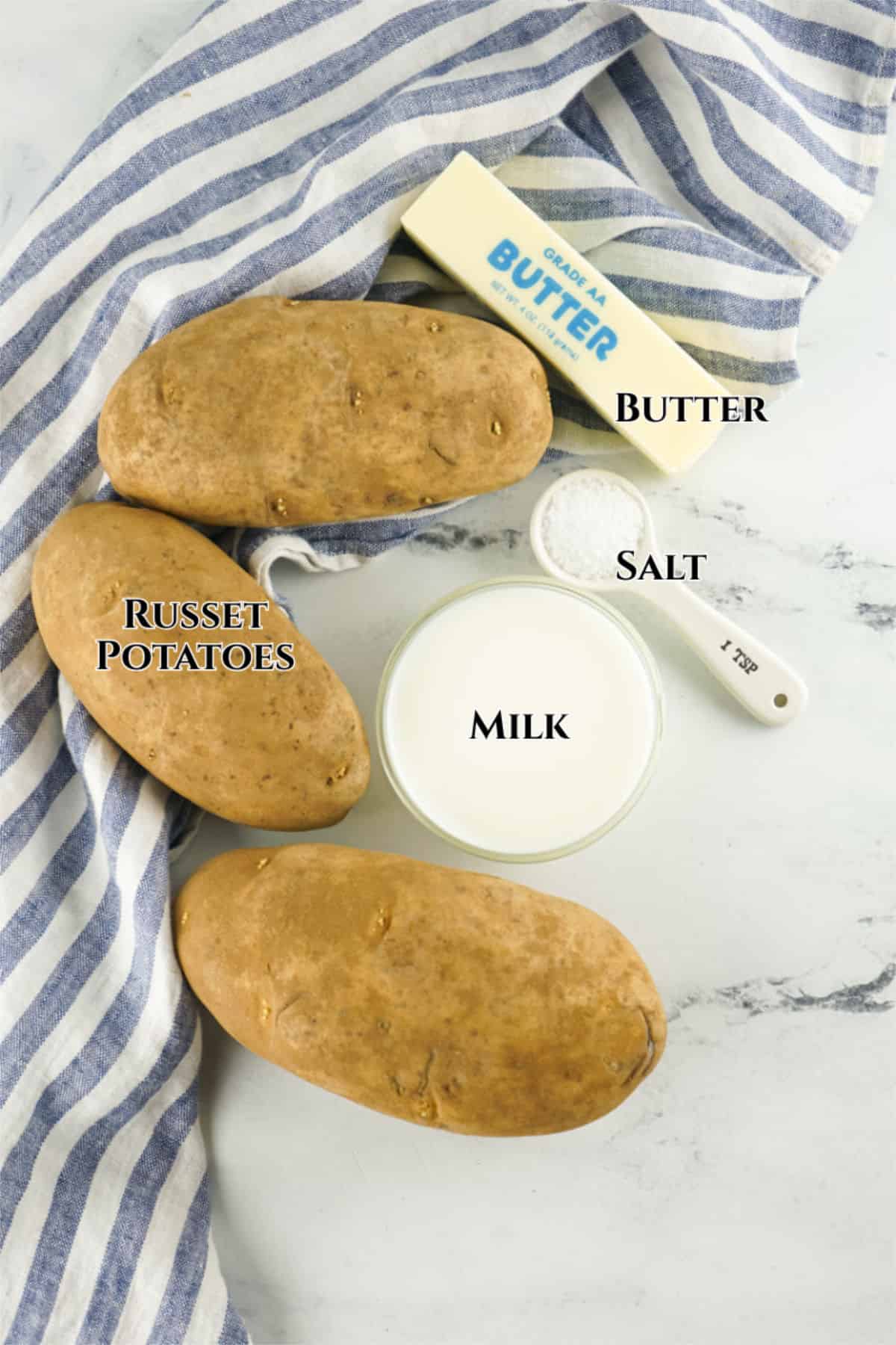 Three russet potatoes, a stick of butter, a small bowl of milk, and a measuring spoon of salt are arranged on a white marble surface with a blue-and-white striped cloth—perfect for making Bob Evans mashed potatoes. Each ingredient is labeled with text.