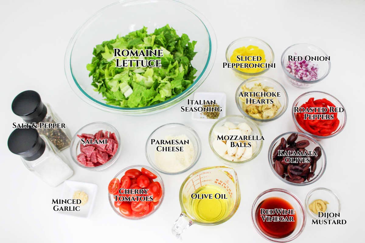 Top view of chopped antipasto salad ingredients in bowls on a white surface, featuring romaine lettuce, sliced pepperoncini, red onion, artichoke hearts, roasted red peppers, salami, parmesan, mozzarella balls, olives, tomatoes, and seasonings.