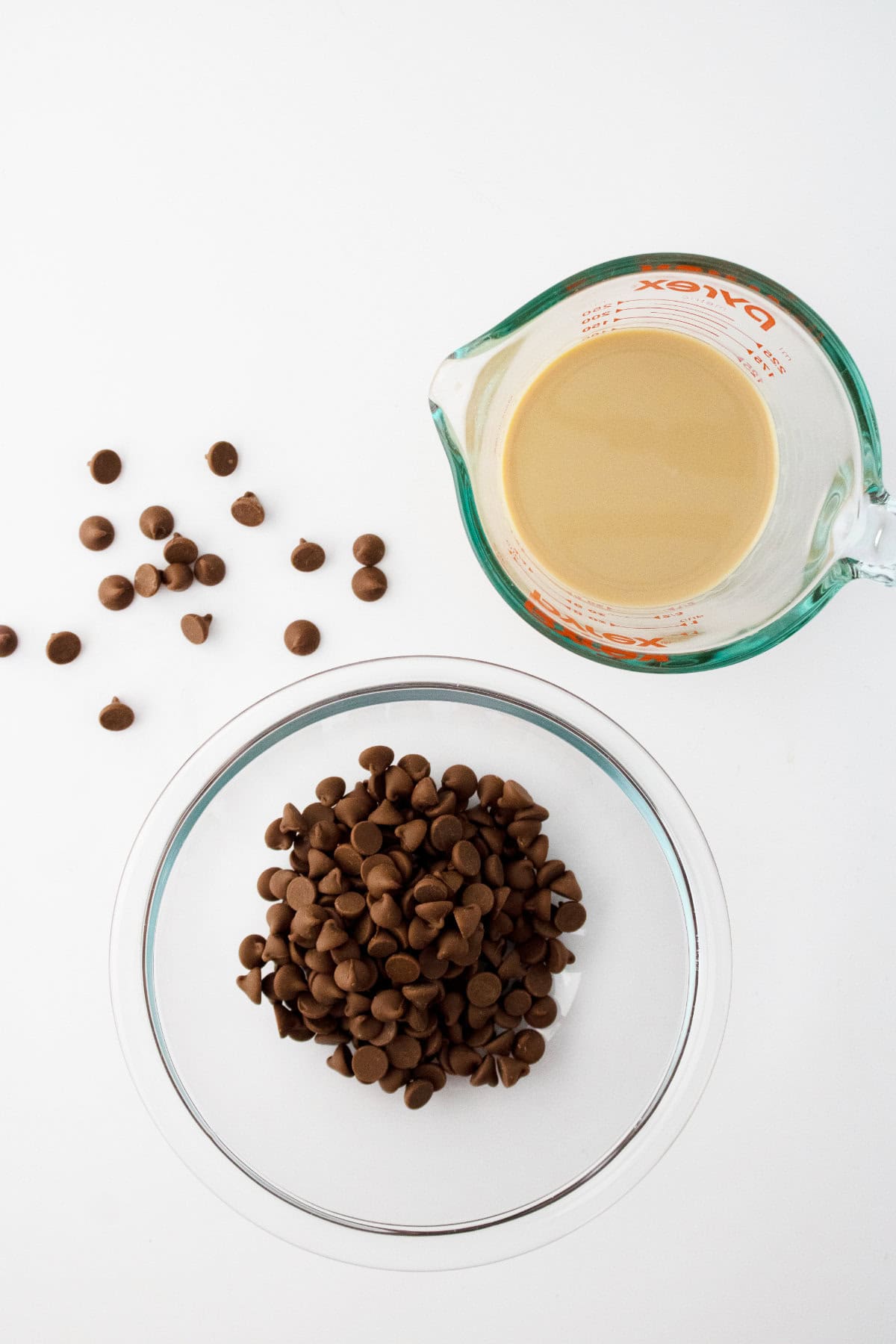 A glass measuring cup with light brown Baileys chocolate ganache and a clear bowl filled with chocolate chips sit on a white surface, with additional chocolate chips scattered nearby.