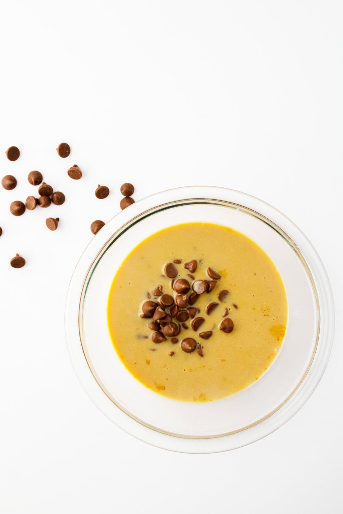 A bowl of creamy yellow soup topped with Baileys chocolate ganache chips, with extra chocolate chips scattered on the white surface beside the bowl.