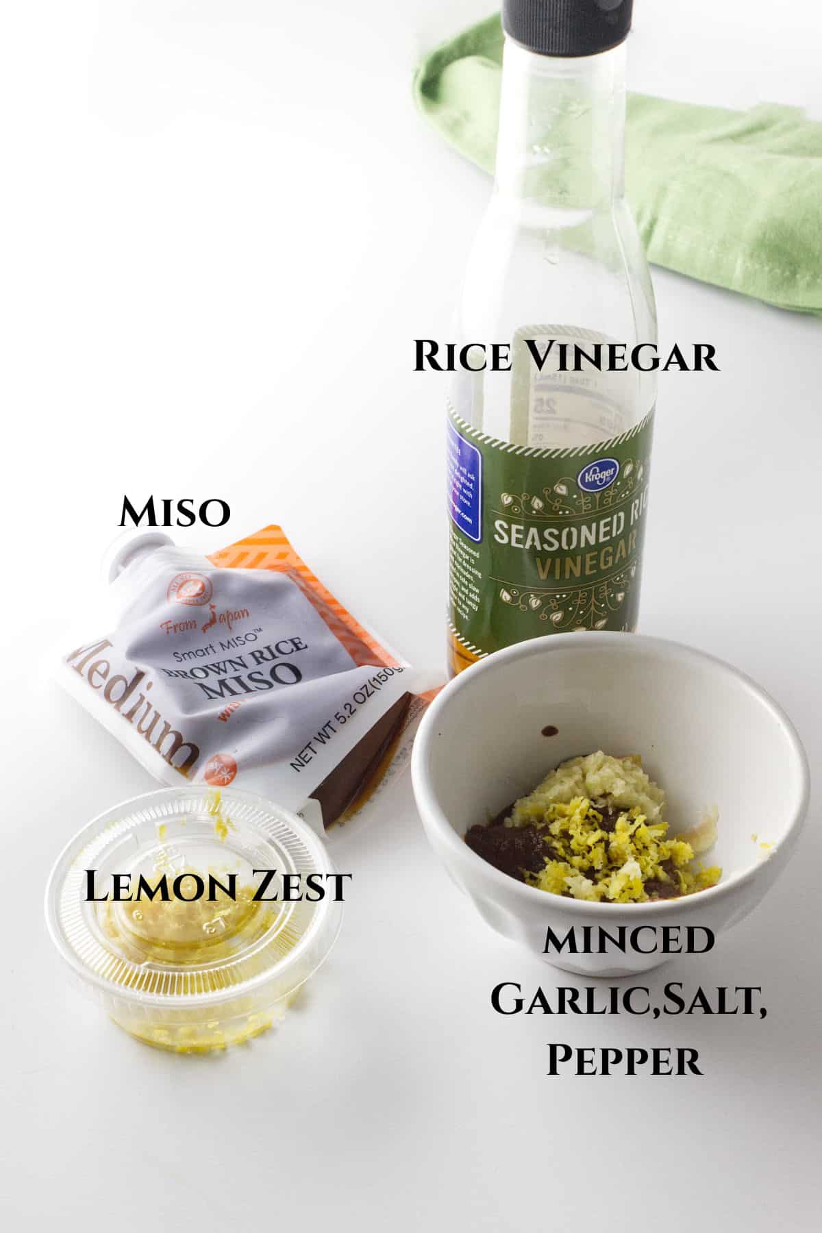 A bottle of rice vinegar, a packet of miso, a small container of lemon zest, and a bowl with minced garlic, salt, pepper, and other ingredients on a white surface. Labels identify each ingredient.