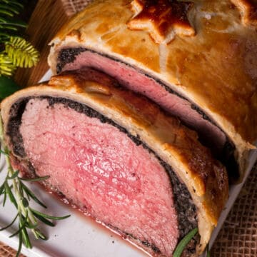 A close-up of beef Wellington shows medium-rare beef wrapped in golden puff pastry, savory mushroom duxelles, and a garnish of rosemary on a white plate—a classic among beef recipes.