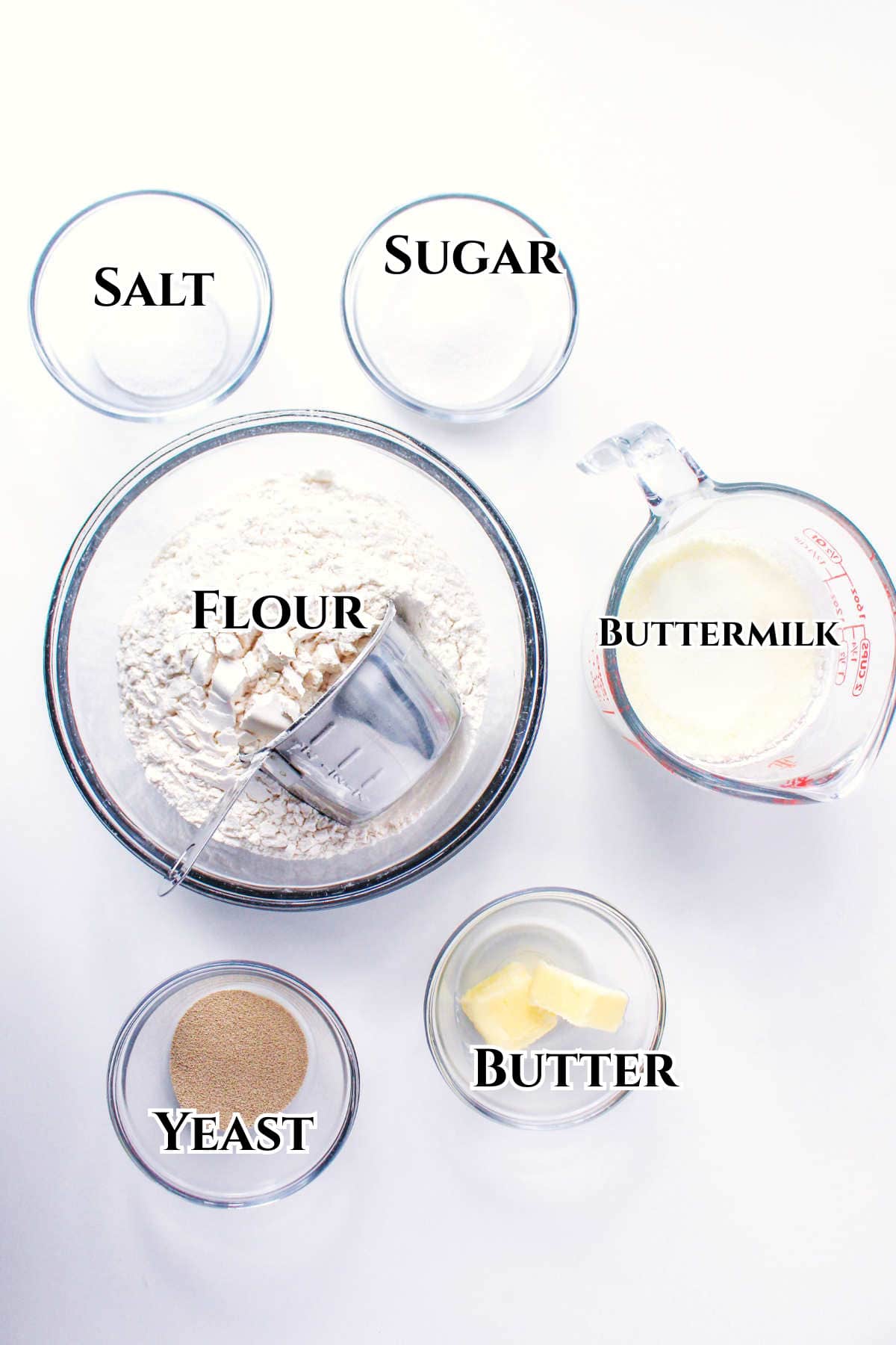 Six glass bowls and a measuring cup containing salt, sugar, flour, buttermilk, yeast, and butter—essential ingredients for bread machine sandwich bread—are arranged on a white surface, each labeled with its ingredient name.