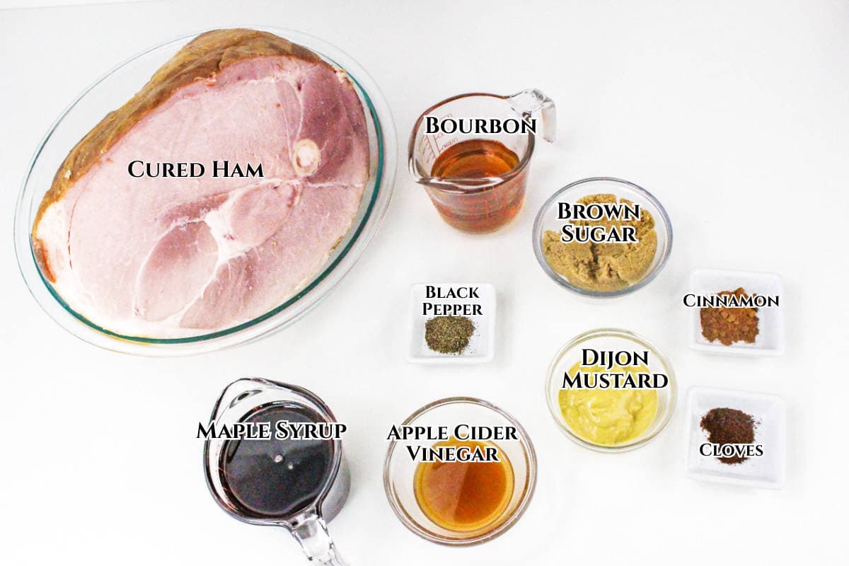 A glass dish with a large bourbon maple glazed ham, and small containers of bourbon, brown sugar, black pepper, cinnamon, cloves, Dijon mustard, maple syrup, and apple cider vinegar arranged on a white surface.