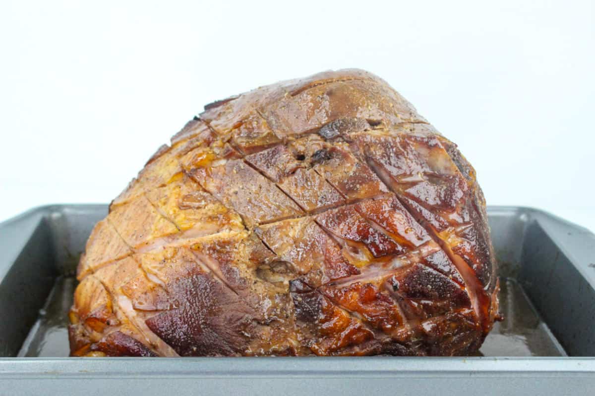 A whole bourbon maple glazed ham with a browned, glossy surface and a crisscross pattern sits in a gray roasting pan against a plain white background.